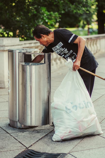 A middle-aged man with dark hair, wearing a black T-shirt with white text and black trousers, is bending over a cylindrical stainless steel outdoor rubbish bin located on a paved pathway in a park or garden setting. The bin has a polished, reflective surface and a rectangular opening at the top. The man is holding a large white refuse sack filled with rubbish in his left hand, while his right hand is reaching into the bin, possibly to dispose of litter or clear it out. The background features lush green trees and a stone balustrade, indicating a well-maintained public area. Natural daylight illuminates the scene, highlighting the metallic finish of the waste receptacle and the vibrant greenery. The scene exemplifies independent waste disposal activity, suggesting support for private rubbish removal services provided by companies like Rubbish Removal Hampstead in the context of alternative waste handling or on-site clearance services.