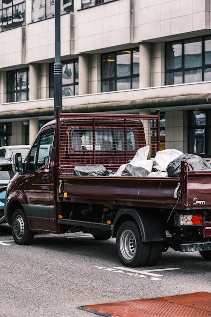 A maroon flatbed pickup truck parked on an urban street in front of a modern, multi-story building with large rectangular windows and light-colored stone cladding. The truck's open cargo bed is filled with various types of rubbish, including large black and grey plastic bags, some of which appear to contain household waste. The metal side and rear barriers of the cargo area are visible, with the barrier at the front of the bed made of a red metal grid for safety and containment. The truck's rear wheel and part of the rear bumper are also visible, and the vehicle is positioned next to a grey marking on the road, possibly indicating a loading or parking zone. The scene conveys an instance of private rubbish removal, typical of independent waste collection services like those provided by Rubbish Removal Hampstead, especially in the context of on-site clearance or alternative waste handling beyond standard local authority collection.