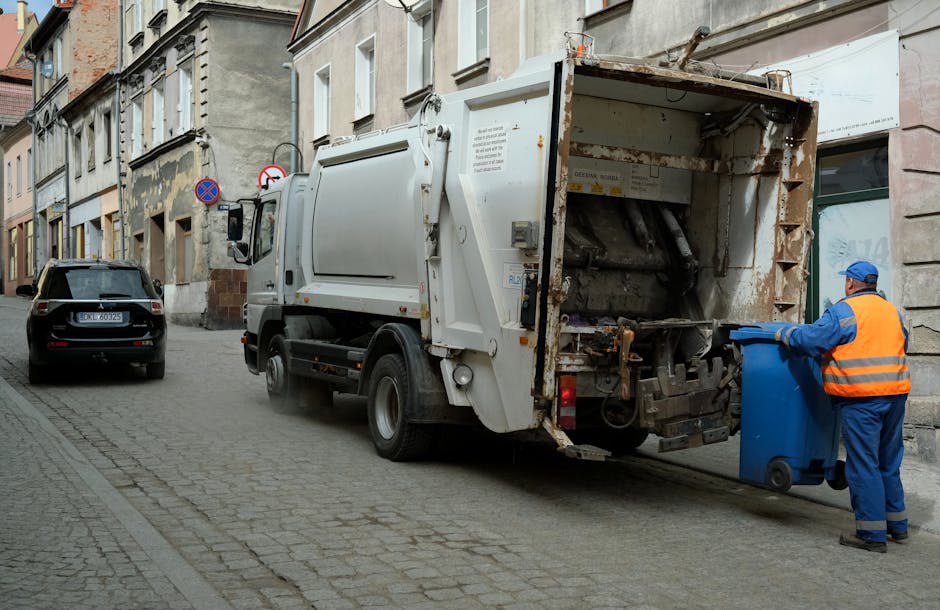 A sanitation worker dressed in blue coveralls and wearing an orange high-visibility vest is shown placing a blue recycling bin into the rear compartment of a white waste collection truck on a cobblestone street. The truck, parked alongside a row of old, weathered residential buildings with multiple floors, has a rusty, utilitarian appearance with signs of wear on its metal exterior. The open rear hopper reveals mechanical components used for compacting rubbish. In the background, there is a black car parked close to the curb, and a 'no parking' sign affixed to a pole on the street. The scene is lit by natural daylight, providing clear visibility of textures such as the smooth finish of the bins, the rough, aged brickwork of the buildings, and the uneven cobblestones underfoot. This image illustrates an example of private waste collection, supported by waste management services like Rubbish Removal Hampstead, emphasizing on-site or alternative rubbish disposal methods outside standard council collection.