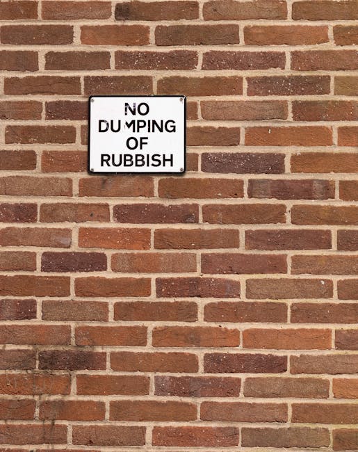A close-up view of a brick wall with a metal street sign attached, reading 'LOWER TERRACE NW3' in uppercase white letters on a green background. The wall is composed of reddish-brown bricks with a rough, textured surface, laid in a traditional pattern with dark mortar joints. The sign is positioned towards the left side of the image, slightly tilted, and appears to be weathered. In the background, beyond the wall, there is a blurred, natural outdoor environment featuring greenery and trees, with soft, diffused light suggesting an overcast day or shaded setting. The image captures an urban scene that could relate to a residential or service area, such as rubbish removal, situated along an alley or pathway adjacent to the brick building. The overall composition emphasizes the street name sign as the primary object within a quiet, suburban context indicating an address or location relevant to private waste handling or local disposal services, such as those offered by Rubbish Removal Hampstead.