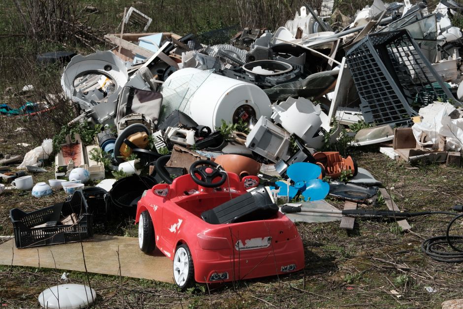 The image depicts a large, disorganized pile of assorted household waste and discarded items scattered outdoors on a patch of grass and dirt. Prominent in the foreground is a small red plastic children's ride-on car with black details, showing signs of wear such as chipped paint. Surrounding the car are various smaller items including a black plastic crate, a white ceiling light fixture, and a tangled black cable. In the background, the heap contains a mixture of broken appliances, plastic containers, and household objects, including a white washing machine drum, a black grill panel, and a variety of white, beige, and grey plastic parts. The scene appears to be an unmanaged waste dump, with items stacked and sprawled randomly, partly surrounded by sparse vegetation and some trees, indicating an outdoor environment. The lighting is natural, casting soft shadows, emphasizing the clutter and disorder of the waste, which could be collected or removed by independent waste clearance services such as Rubbish Removal Hampstead, especially when dealing with bulky or mixed household rubbish.