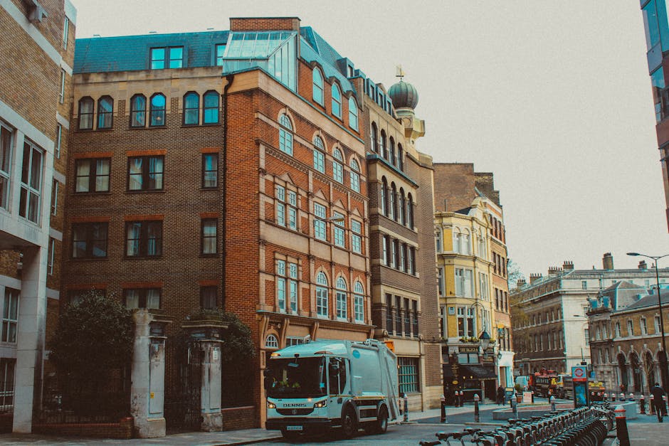 A blue and white waste collection truck is parked on a city street surrounded by multi-story buildings featuring a mix of brick and ornate architectural details, including arched windows and decorative facades. The truck, positioned towards the foreground on the left side, has a closed rear compartment used for rubbish collection, with visible side mirrors and a cab with dark-tinted windows. Adjacent to the truck, the street is lined with black metal bollards and bicycle racks, with a sidewalk that has minimal pedestrian activity. The environment appears urban, with the background showing other vehicles and historic-style buildings, some with domed rooftops and decorative spires. The scene takes place in daylight under an overcast sky, indicating typical city waste management activity, potentially reflecting an alternative to on-site rubbish disposal or private waste collection services. The presence of Rubbish Removal Hampstead is implied through the context of waste collection, highlighting professional rubbish removal in the area.