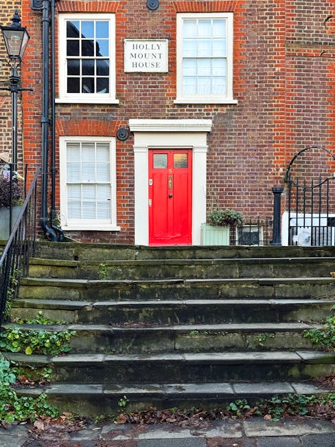 The image shows the front facade of a three-story brick building with a red wooden front door situated at the top of a short flight of stone steps. The steps appear weathered with patches of moss and small plants growing between the cracks, indicating age and outdoor exposure. To the left of the door, there is a small green potted shrub and possibly a decorative planter, adding a touch of greenery to the entrance. The building features white-framed sash windows, with two directly above the door and one at ground level, all with white blinds or curtains. A sign reading 'HOLLY MOUNT HOUSE' is mounted on the brick wall between the upper windows. Iron railings run along the sides of the stairs, with a black street lamp visible on the left. The overall scene suggests an older residential property in an urban setting, with natural light illuminating the building's textured brickwork, emphasizing the weathered stones and brick details. The image context aligns with independent waste management or local authority alternative collection options as part of rubbish removal services offered by Rubbish Removal Hampstead.