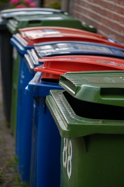 A row of large rigid wheelie bins lined up along the edge of a pavement adjacent to a red brick wall. The bins are predominantly blue, with green, red, and dark green lids visible, some slightly open revealing the textured surfaces of the lids. The bins are made of durable plastic with a slightly glossy finish, and the lids feature ribbed patterns for grip. The scene is outdoors, on a residential street, with natural daylight illuminating the bins and casting subtle shadows. The background shows the continuation of the brick wall and a glimpse of an overcast sky. This arrangement of bins is typical for private waste collection, as managed by waste removal services such as Rubbish Removal Hampstead, providing an alternative to municipal rubbish disposal, often used for on-site clearance or private collection needs in the area.