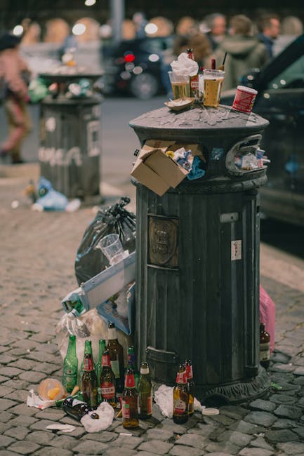 A black, cylindrical street trash bin made of metal with a domed lid is shown on a cobbled pavement, surrounded by scattered trash including glass bottles, paper, and plastic waste, indicating recent littering. The lid of the bin has several items placed on top, such as a disposable coffee cup with a plastic lid, a paper food container, and a small cardboard box. To the left of the bin, a black garbage bag is partially visible, bulging with waste. The background features a blurred urban scene with a grey car parked nearby and pedestrians walking along the street. The environment suggests an outdoor public space where rubbish has accumulated and discarded items are visible, emphasizing the importance of rubbish removal and proper waste disposal. The scene might be associated with discussions of independent waste collection or on-site rubbish clearance in urban areas, as highlighted by the presence of local waste management services like Rubbish Removal Hampstead. The overall atmosphere appears typical of city street cleanliness challenges, with a focus on maintaining tidy public spaces through appropriate rubbish management.