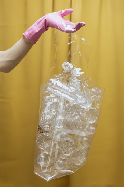 A close-up view of a person wearing a pink glove, holding a clear plastic bag filled with crushed and whole transparent plastic bottles, with a yellow curtain in the background. The bottles are made of PET material with smooth, shiny surfaces, and vary slightly in size and shape. The bag is tightly filled and appears to be prepared for recycling or disposal, illustrating a typical scene of waste collection or private rubbish handling. The environment suggests an indoor area or a designated waste collection space, relevant to rubbish removal services such as those offered by Rubbish Removal Hampstead, which often includes handling plastics as part of sustainable waste management practices.