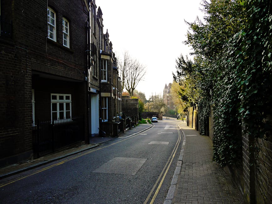 A narrow residential street showing a row of brick-built terraced houses on the left side, with black metal railings and white-framed windows. The street surface is asphalt with visible double yellow lines along the curb, indicating parking restrictions. The right side features a paved pedestrian walkway bordered by a high, dense hedge of green foliage, with some branches extending over the pavement. In the background, leafless and partly leafed trees are visible, along with a distant building that appears to be a church or institutional structure with a pointed roof and tall windows. The scene is illuminated by natural daylight, with a softly overcast sky, and no visible waste or rubbish in the image. This setting, by showcasing a tidy street environment, subtly relates to the services of private rubbish removal and on-site clearance, as provided by Rubbish Removal Hampstead, somewhere where independent waste handling may complement local authority services.