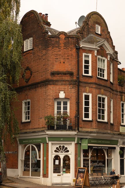 The image depicts a three-story corner building constructed with red brick in a traditional architectural style, featuring decorative brickwork and a curved parapet roof. The ground floor functions as a commercial space with a green and white facade, including large display windows and a central entrance door framed by decorative white trim and a fanlight window. A small black chalkboard sign is placed outside on the pavement, indicating it may be a cafe or similar establishment. Above the shopfront, three floors of residential windows are visible; the first and second floors have white-framed sash windows, with the second floor featuring a small balcony adorned with potted plants and flowers. The building is situated on a street corner with a sidewalk, and a large leafy tree partially obscures the left side of the scene. The sky is overcast, and the area appears to be a busy urban setting. The scene subtly evokes the context of private or independent waste handling in commercial and residential premises, such as collection or clearance services by Rubbish Removal Hampstead.