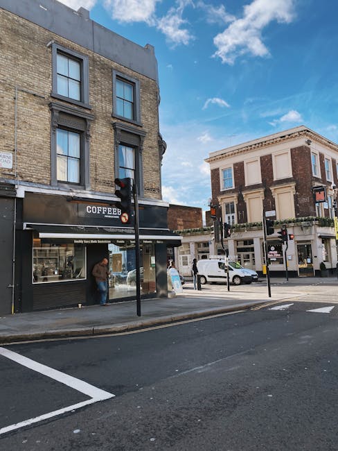 A street view in Hampstead showing the ground floor of a three-story brick building with large, rectangular windows and dark trim, housing a coffee shop with a black sign that reads 'COFFEE' in white letters. A person wearing a brown jacket and blue jeans stands near the shop's window, possibly ordering or waiting. To the right, there is a two-story corner building with a cream-colored facade, decorative molding, and large windows on both levels, featuring signs and awnings indicating commercial premises. A white van is parked along the curb behind a row of black bollards, and several pedestrians are visible crossing or waiting at the crosswalk. The scene is illuminated by natural daylight, with a partly cloudy sky overhead, and the surrounding environment includes asphalt roads and sidewalks, with some street furniture such as traffic lights and posts. The image is related to local waste management and suggests the importance of on-site clearance or private disposal practices supported by local rubbish removal services like Rubbish Removal Hampstead.