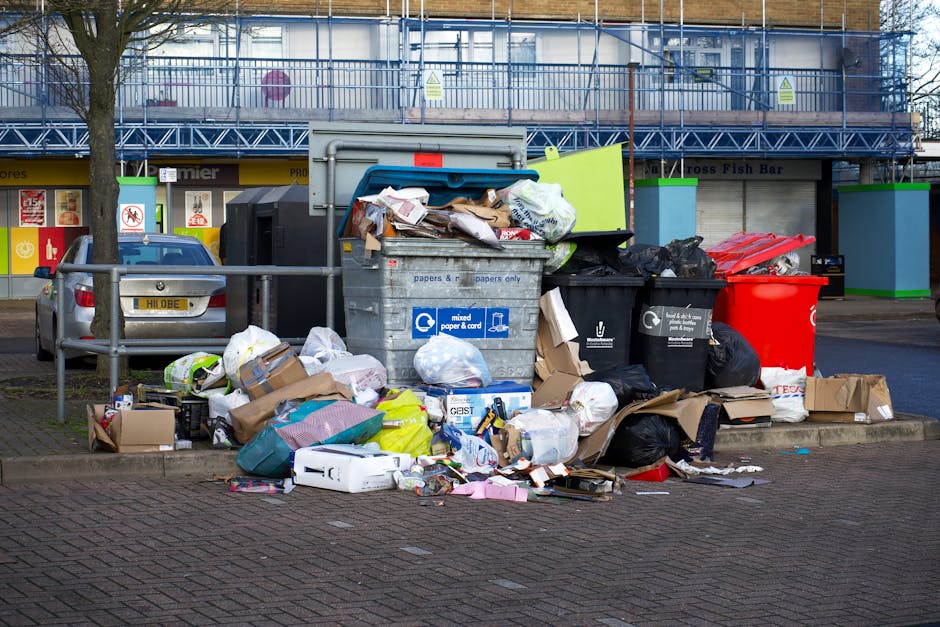 A large collection of overflowing rubbish bins and numerous scattered waste items on a paved street in front of a commercial building, with some black and red wheelie bins opened and filled with mixed waste, including cardboard boxes, plastic bags, paper, and packaging materials. The scene shows various types of waste materials piled on the ground, with some items spilling out from open bins. A silver car is parked nearby, with a tree and a metal railing separating the waste area from the adjacent sidewalk. In the background, a building under construction or renovation is visible, covered with blue scaffolding and protective sheeting, alongside shopfronts with closed shutters. The outdoor scene is illuminated by daylight, creating a contrast between the waste contamination and the surrounding environment, and highlighting the importance of proper rubbish removal, as provided by services such as Rubbish Removal Hampstead.