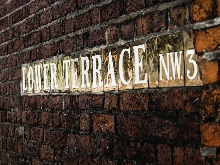 A close-up view of a rectangular ceramic street sign mounted on a weathered red brick wall, displaying the text 'LOWER TERRACE N.W.3' in large, white, capital letters with a slightly distressed appearance. The sign's surface shows signs of corrosion and moss growth, particularly around the edges and on the white lettering, indicating outdoor exposure. The bricks beneath are arranged in a horizontal pattern with visible mortar joints, and the wall has a textured, aged look with variations in color from deep red to darker shades. The lighting highlights the uneven surface of the brickwork and the reflective qualities of the glazed sign, creating a subtle contrast between the darkened areas and the brighter illuminated parts of the sign. This scene suggests an urban setting in a historical or older residential area, fitting with the context of private waste handling and alternative collection methods by local waste management services like Rubbish Removal Hampstead.
