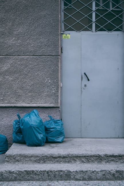 A large collection of overflowing rubbish bins and numerous scattered waste items on a paved street in front of a commercial building, with some black and red wheelie bins opened and filled with mixed waste, including cardboard boxes, plastic bags, paper, and packaging materials. The scene shows various types of waste materials piled on the ground, with some items spilling out from open bins. A silver car is parked nearby, with a tree and a metal railing separating the waste area from the adjacent sidewalk. In the background, a building under construction or renovation is visible, covered with blue scaffolding and protective sheeting, alongside shopfronts with closed shutters. The outdoor scene is illuminated by daylight, creating a contrast between the waste contamination and the surrounding environment, and highlighting the importance of proper rubbish removal, as provided by services such as Rubbish Removal Hampstead.